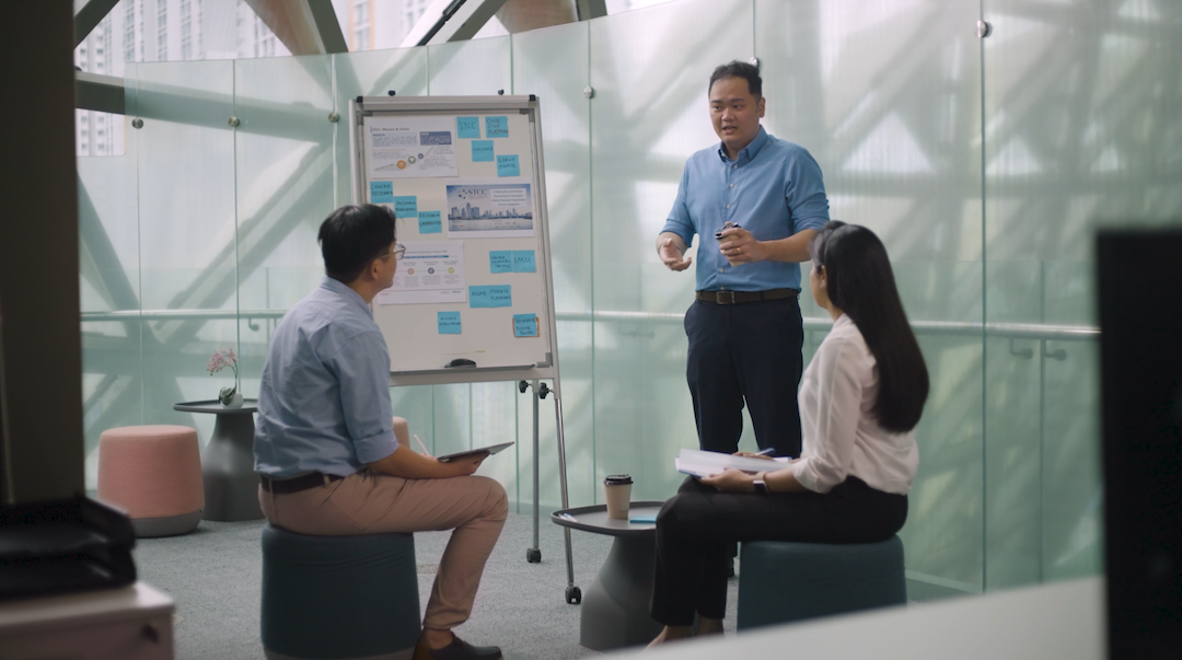Three people sit and listen to a presentation in a modern office space with a flip chart displaying graphs and post-it notes.